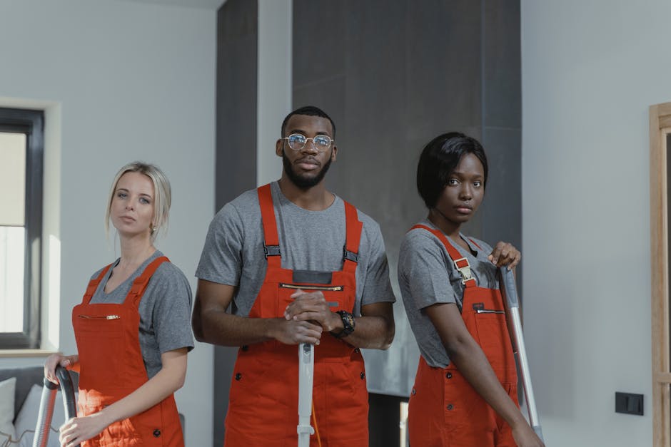 Three cleaning professionals standing indoors, dressed in grey t-shirts and orange aprons, each holding cleaning equipment such as vacuum cleaner and mop. The background features a modern, well-lit ro