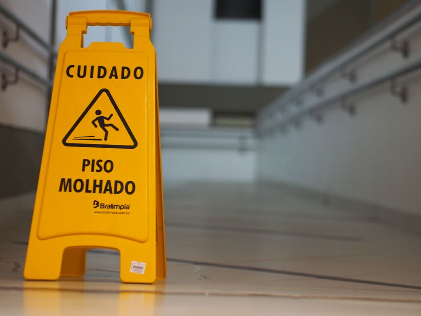 Three cleaning professionals standing indoors, dressed in grey t-shirts and orange aprons, each holding cleaning equipment such as vacuum cleaner and mop. The background features a modern, well-lit ro