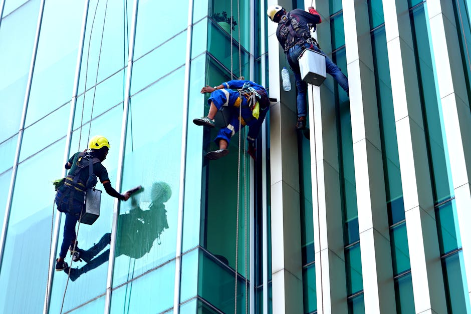 Three professional window cleaners wearing yellow safety helmets and harnesses are performing surface cleaning and window washing on the exterior glass facade of a modern office building. They are usi